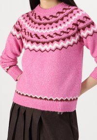 Rosa stickad tröja med rund halsringning, prydd med ett geometriskt mönster i brunt och vitt vid kragen och nederkanten. Mjuk textur, avslappnad passform.