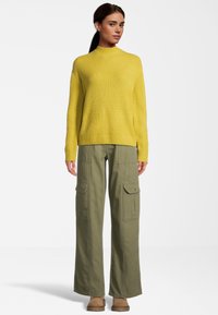 Gelber gerippter Strickpullover mit hohem Kragen, kombiniert mit weit geschnittenen olivgrünen Cargo-Hosen, die Seitentaschen und eine lockere Passform aufweisen.