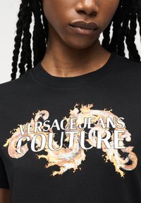 Γυναίκα που φοράει μαύρο μπλουζάκι με το λογότυπο "Versace Jeans Couture" και διακοσμητικά χρυσά στροβιλιζόμενα σχέδια στο στήθος.
