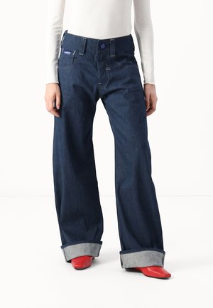 Person iført mørkeblå jeans med vide ben og store manchetter samt klare røde spidse støvler, kombineret med en hvid langærmet top.