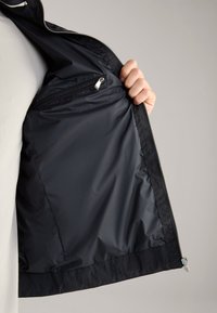 Hand die een open zwart jasje vasthoudt, waarbij de gladde binnenvoering en een klein zakje met rits zichtbaar zijn, tegen een lichtgrijze achtergrond.