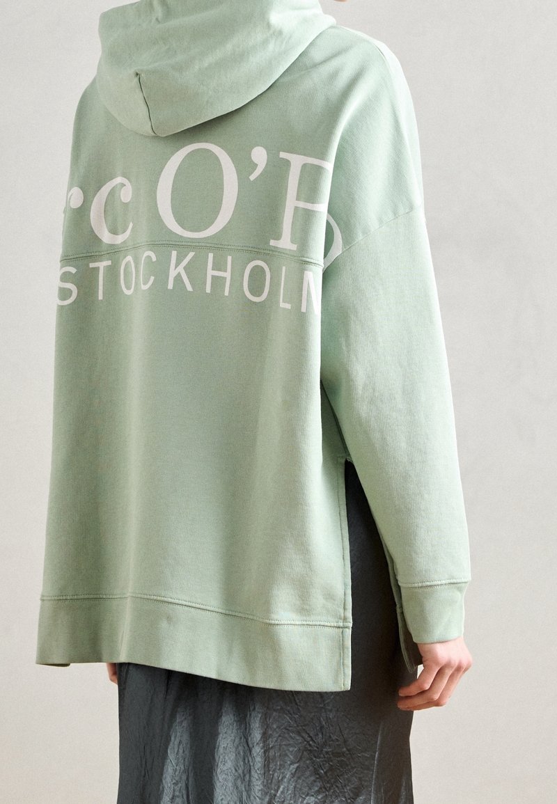 Personne portant un sweat à capuche ample vert clair avec « STOCKHOLM » imprimé en blanc et une jupe sombre, photographiée de dos sur un fond uni.