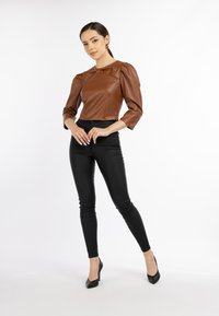 Braune Lederjacke im Cropped-Stil mit Puffärmeln, kombiniert mit schwarzen, eng anliegenden Hosen und schwarzen High Heels, vor einem neutralen Hintergrund.