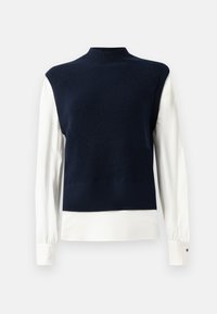 Marineblaue ärmellose Strickweste, die über einer weißen Bluse mit langen Ärmeln und Knopfmanschetten auf einem schlichten Hintergrund getragen wird.