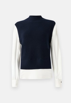Marineblauwe mouwloze gebreide vest over een witte blouse met lange mouwen en knoopmanchetten, tegen een effen achtergrond.