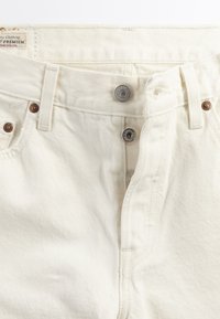 Λευκά τζιν από τζιν ύφασμα με κλείσιμο με ασημένια κουμπιά, θηλιές για ζώνη και χαλκοσωλήνες στις μπροστινές τσέπες, που δείχνουν την υφή του υφάσματος και τις λεπτομέρειες ραφής.