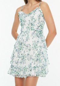 Blumenkleid im Mini-Stil mit verstellbaren Trägern, gerafftem Ausschnitt und geschichteten Saum. Hat einen weißen Hintergrund mit grünen und lila Blumenmustern.