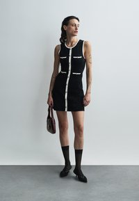 Czarna dopasowana sukienka z białymi guzikami, z okrągłym dekoltem i kieszeniami. W połączeniu z czarnymi skarpetkami i spiczastymi balerinami, trzymająca bordową torebkę.