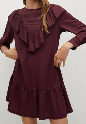 Femme portant une robe prune à manches longues avec un volant en forme de V sur la poitrine et un ourlet froncé, ajustant les coutures latérales.