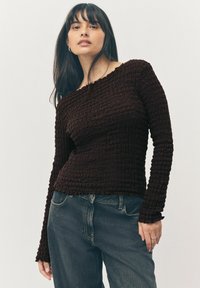 Hnědý texturovaný top s dlouhým rukávem a žebrovaným designem, s výstřihem na ramena a přiléhavým siluetou, zkombinovaný s tmavými denimovými džíny.