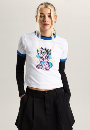 Jonge vrouw met blond haar in laagjes draagt een wit cropped T-shirt met blauwe rand en een cartoon puppy-afbeelding, zwarte lange mouwen en een zwarte rok.