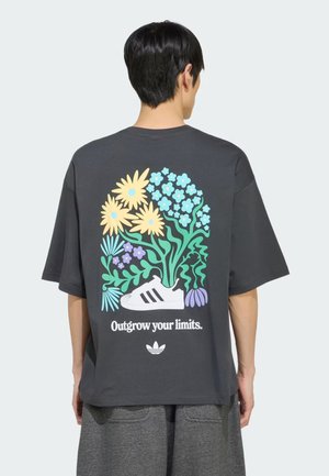 Person trägt dunkelgraues T-Shirt mit buntem Blumenmotiv und weißer Sneaker-Grafik, Text "Outgrow your limits" auf dem Rücken.