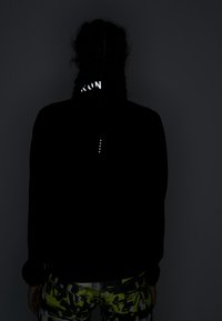 Henkilö, jolla on letitetyt hiukset, kääntynyt poispäin, pukeutuneena mustaan takkiin, jossa on heijastava "RUN"-teksti kauluksessa ja kuvioituun kelta-mustaan housupariin.