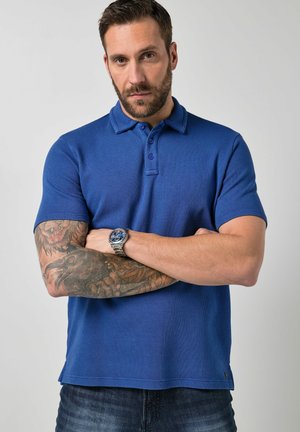 Mężczyzna z brodą i skrzyżowanymi ramionami, ubrany w niebieską koszulkę polo i zegarek na nadgarstku, prezentujący szczegółowe tatuaże na lewej przedramieniu i ramieniu.
