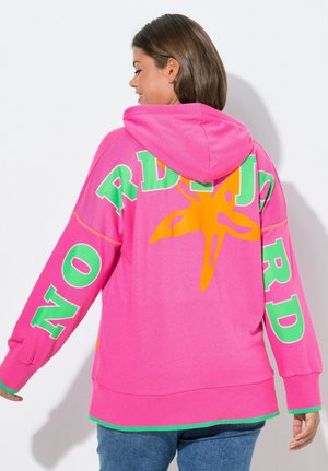 Frau trägt leuchtend pinken Hoodie mit grünen Buchstaben und einem großen orangen Seestern-Design auf dem Rücken, kombiniert mit blauen Jeans.
