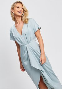 Svetlo modra obleka iz mehkega blaga, z globokim V-izrezom, kratkimi rokavi in asimetričnim robom z drapiranjem.