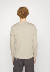 Pitkähiha beige paita, joka on valmistettu pehmeästä puuvillasta. Tässä paidassa on pyöreä kaula-aukko ja yksi hihapokka. Pintakuviointi on teksturoitu ja pinta sileä.