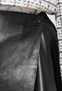 Primo piano di un indumento in pelle nera con un bottone nero, sovrapposto a un maglione crema e a un tessuto intrecciato bianco e blu.