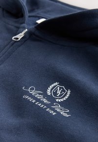 Sweat à capuche zippé bleu marine avec texture douce, arborant un logo blanc avec le texte "Upper East Side" et des accents de laurier décoratifs.