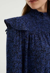 Žena v modré blůze s černými abstraktními skvrnami, s nařaseným výstřihem a volánkovými detaily na rameni, zobrazená od krku až po horní část hrudníku.