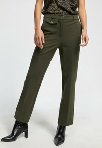 Pantalones de corte entallado en verde oliva con una textura suave, que cuentan con bolsillos delanteros y cierre de botón, combinados con tacones negros de punta.