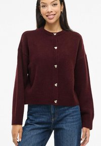 Bordeauxroter Strickpullover mit rundem Ausschnitt und oversized Schnitt, ausgestattet mit sechs herzförmigen goldenen Knöpfen vorne. Strukturiertes Gewebe.