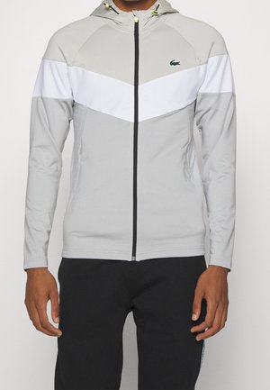 Mies, jolla on vaaleanharmaa vetoketjullinen huppari, jossa on valkoinen V-kirjaimen muotoinen rintaraita ja pieni vihreä krokotiililogo vasemmalla puolella.