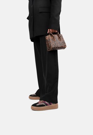 Osoba w czarnym garniturze trzymająca małą brązową torebkę w panterkę, nosząca czarne trampki z wzorem w panterkę i różowymi akcentami.