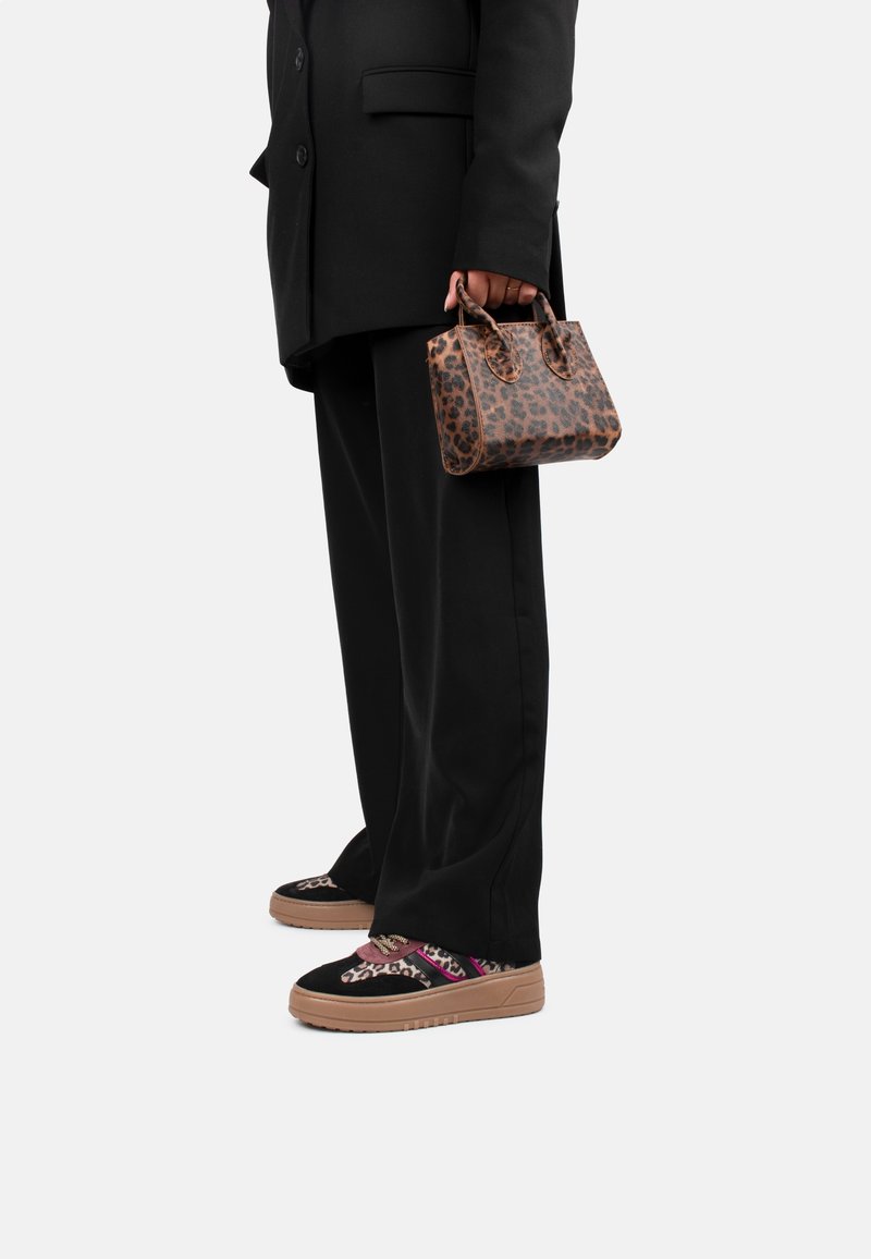 Personne en costume noir tenant un petit sac à main marron à motif léopard, portant des baskets noires avec motif léopard et accents roses.