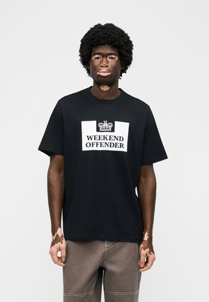 Άνδρας με λεύκη που φοράει μαύρο t-shirt "Weekend Offender" και γκρι παντελόνι, στέκεται μπροστά σε απλό ανοιχτό φόντο.