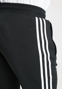 Svarta sweatpants gjorda av mjukt tyg har en bred elastisk midja och tre vertikala vita ränder längs sidan.