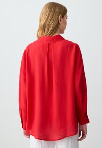 Red long-sleeve shirt made of lightweight fabric, featuring a structured collar, back pleat, and buttoned cuffs. Smooth texture with a relaxed fit.