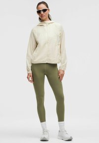 Creme vindjakke med hætte, olivenfarvede leggings og hvide sneakers. Slankt design med lange ærmer og lynlås foran.