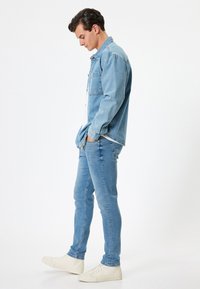 Giacca di jeans azzurro chiaro con una sola tasca sul petto, abbinata a jeans attillati azzurro chiaro e sneakers bianche, in piedi su uno sfondo semplice.