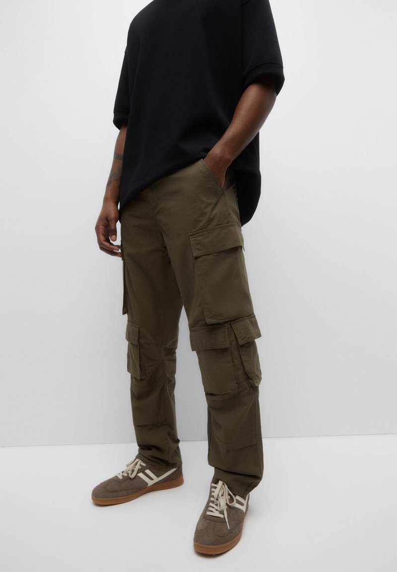 Pantalon cargo vert olive avec multiples poches, coupe droite et tissu robuste. Associé à des baskets marron et un t-shirt noir oversize.