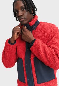 Czerwony polarowy kurtka z teksturowaną powierzchnią, granatowymi akcentami, zatrzaskami i kieszeniami na zamek, z wysokim kołnierzem i luźnym krojem.