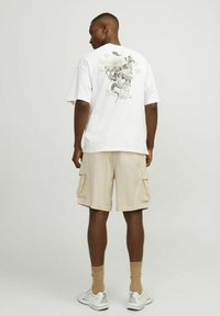 T-shirt blanche surdimensionnée avec un design graphique représentant un crâne et un serpent. Associée à un short cargo beige et des baskets blanches.