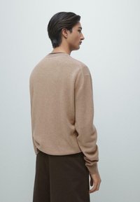 Beige tröja i ett mjukt, texturerat tyg med rund halsringning och ribbade muddar, kombinerad med mörkbruna byxor.