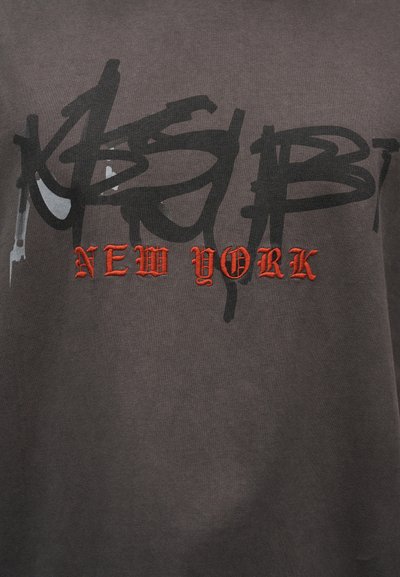 Γκρι βαμβακερό T-shirt με μαύρα γράμματα σε στυλ γκράφιτι και κόκκινα κεντημένα γράμματα που γράφουν «NEW YORK» στο κέντρο.