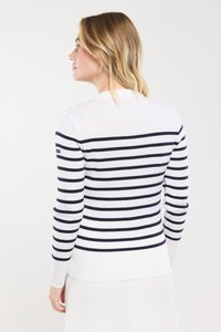 Vrouw met schouderlang blond haar, gekleed in een witte sweater met zwarte horizontale strepen, met de rug naar de camera gekeerd tegen een effen achtergrond.