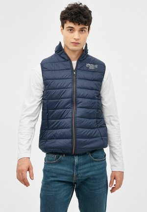 Chaleco tipo plumas en color azul marino con diseño acolchado, cierre de cremallera en la parte delantera y cuello alzado. Presenta un logo en el lado izquierdo del pecho y detalle de cremallera en contraste.
