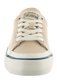 Zapatilla de lona beige con cordones blancos, suela de goma y acento azul en la base. Presenta una etiqueta de logo en la lengüeta. Superficie superior texturizada.
