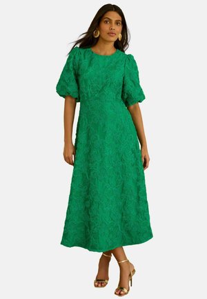 Femme portant une robe midi verte à motifs floraux texturée avec des manches bouffantes et des sandales à talons hauts dorées, debout devant un fond uni.