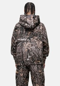 Felpa camouflage con un grande cappuccio e tasca frontale, caratterizzata da una stampa testurizzata di corteccia d'albero e foglie in tonalità terrose di marrone e verde.