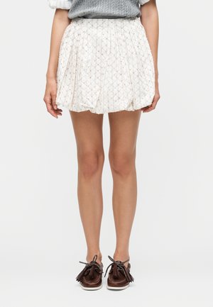 Kobieta ubrana w białą plisowaną spódnicę w wzory, szary sweter i brązowe mokasyny z frędzlami stoi z rękami swobodnie opuszczonymi wzdłuż ciała.