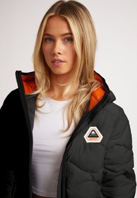 Giubbotto in materiale impermeabile nera con fodera arancione, patch con logo esagonale e texture trapuntata. Caratterizzato da un colletto alto e chiusura con zip.