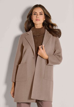 Vrouw met lang bruin haar, gekleed in een beige knielange jas over een bruine turtleneck trui, staand tegen een lichtgrijze achtergrond.