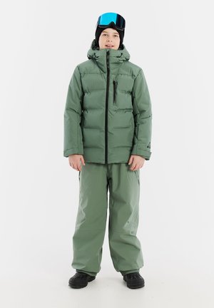 Groene pufferjas met een ritssluiting aan de voorkant, zijzakken en capuchon. Gecombineerd met bijpassende loszittende broek en zwarte laarzen. Blauwe skibril op het hoofd.