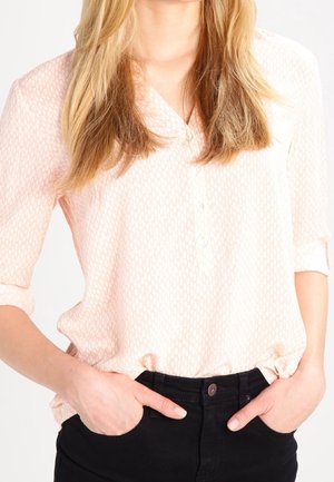 Blouse à boutons rose pâle avec un motif texturé, manches retroussables et coupe décontractée, associée à un pantalon noir, mains dans les poches.