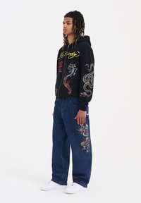 Felpa nera con ricami colorati di drago e serpente; jeans azzurri con design simile sulla gamba; sneakers bianche.